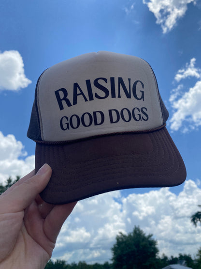 Brown and Tan Foam Trucker Hat with 'Raising Good Dogs' in brown text held against a blue sky with clouds