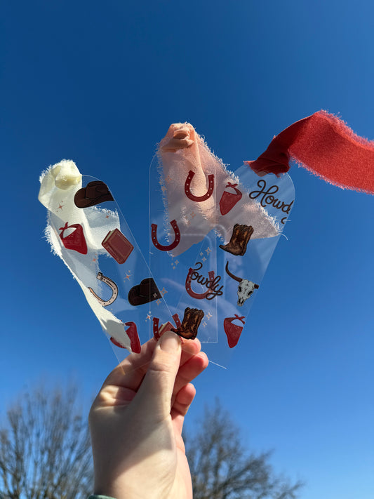 Three cowboy romance acrylic bookmarks with ribbons held against a bright blue sky. The set includes a white ribbon bookmark with a cowboy hat and horseshoe collage, a pink ribbon bookmark with horseshoes and stars, and a dark red ribbon bookmark featuring rope "Howdy" text, a longhorn skull, and cowboy boots. These transparent western page markers are perfect for fans of small-town romance novels and rugged rancher tropes. High-quality clear acrylic gift for bookstagram.