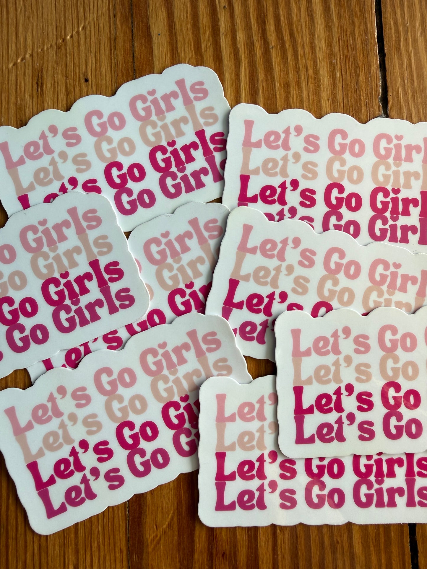 Multiple Let’s Go Girls pink cowgirl stickers laid out on wood surface, western country sticker featuring repeated Let’s Go Girls text in varying pink tones, trendy girly western vinyl stickers for laptop, tumbler, phone case