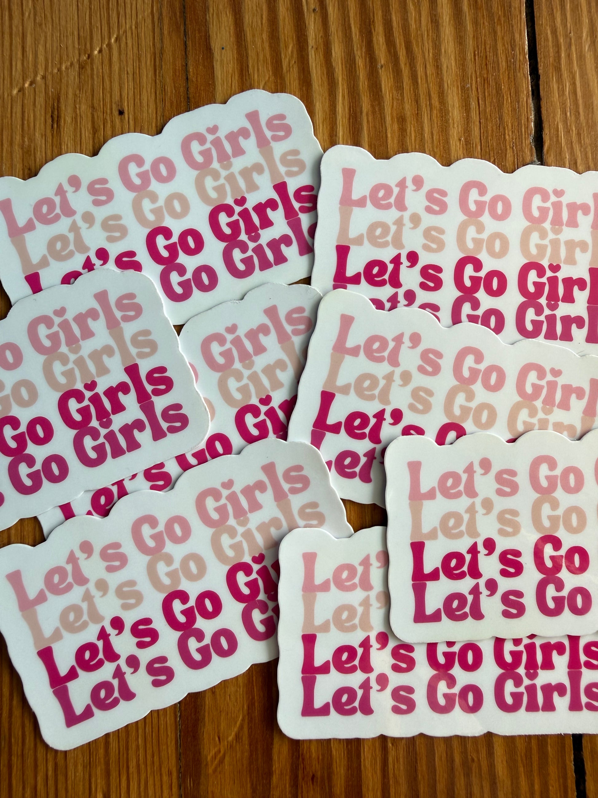 Multiple Let’s Go Girls pink cowgirl stickers laid out on wood surface, western country sticker featuring repeated Let’s Go Girls text in varying pink tones, trendy girly western vinyl stickers for laptop, tumbler, phone case
