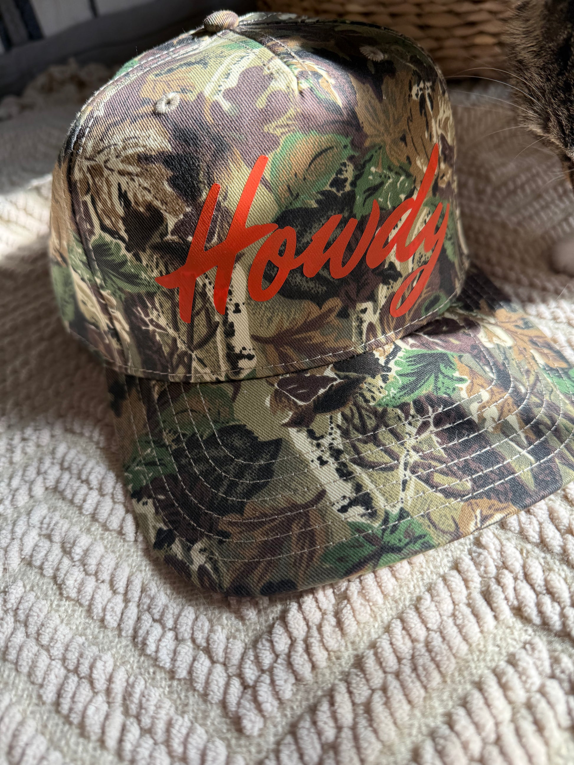 A camouflage baseball cap with orange "Howdy" text resting on a cozy tan knit blanket. Natural sunlight from a nearby window illuminates the fabric texture, highlighting the hat as a perfect gift for country music fans or western lovers.