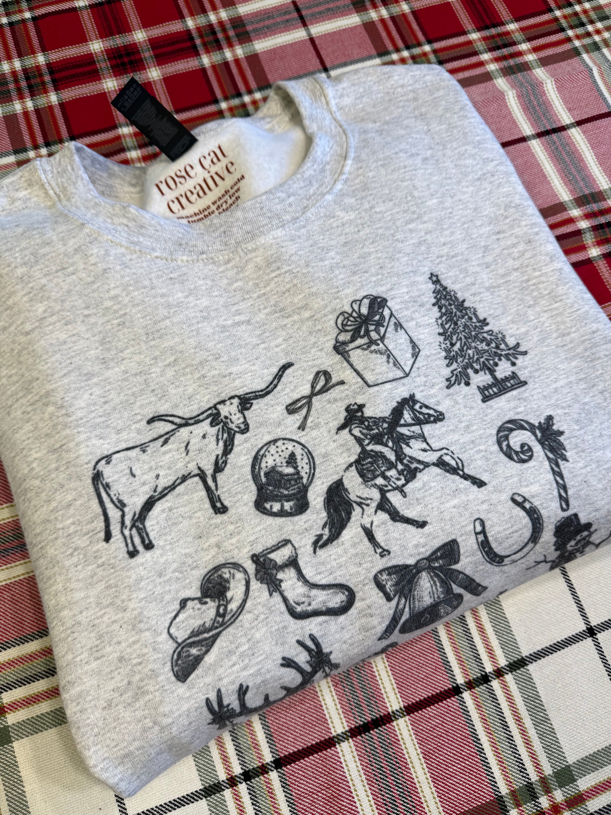 Folded ash-colored cowboy Christmas sweatshirt on a red plaid backdrop, featuring a detailed collage of vintage cowboy art and Christmas illustrations including Western boots, cowboy hats, stars, and holiday accents. Ideal for long-tail Etsy keywords like rustic Western Christmas gift ideas, retro cowboy holiday clothing, and cozy cowgirl aesthetic winter apparel.